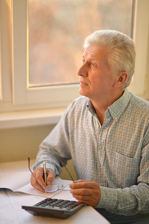 Serious elderly man with calculator sitting at tableの写真素材