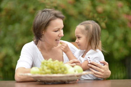 Mother and her little daughter sitting at table outdoors and  fruitsの写真素材