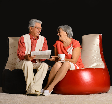 Senior couple reading newspaper with coffee sitting in red armchair on black backgroundの写真素材