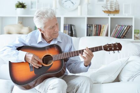 Portrait of happy senior man with guitar sitting on the sofa at homeの写真素材