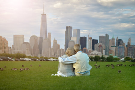 Senior couple sitting on green meadow against New York cityscapeの写真素材