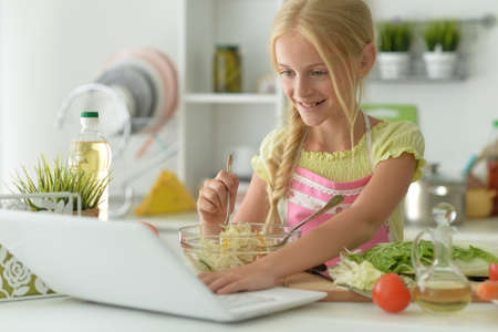 Cute girl making salad on kitchen and using laptopの写真素材
