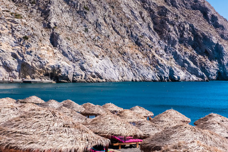 View of beach umbrellas with blue water and a rock in the backgroundの写真素材