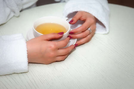 Girl of Slavic appearance in white coat with phone on bed in hotel room. Pretty female shows acting emotions. Young charming woman is sitting on sofa in bedroom and playing with her feelings. Relaxの写真素材