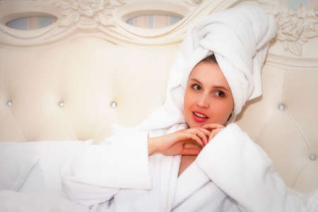 Girl of Slavic appearance in white coat with phone on bed in hotel room. Pretty female shows acting emotions. Young charming woman is sitting on sofa in bedroom and playing with her feelings. Relaxの写真素材