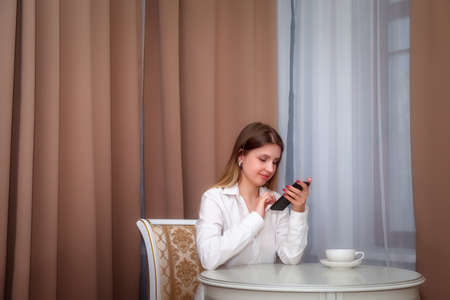 Girl of Slavic appearance in white coat with phone on bed in hotel room. Pretty female shows acting emotions. Young charming woman is sitting on sofa in bedroom and playing with her feelings. Relaxの写真素材