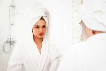 Girl of Slavic appearance in white coat on bed in hotel room. Pretty female shows acting emotions. Young charming woman is sitting on sofa in bedroom and playing with her feelings. Relax conceptの写真素材