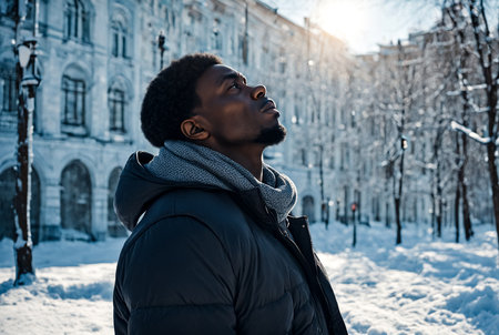 African American man standing in snowy Russian winter at urban background, looking up away. Portrait black man walking of city, falling snow outside. Lifestyle winter concept. Copy ad text spaceの素材