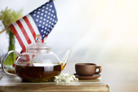 Glass teapot with tea brown cup on a wooden table. American flag in the background. Festive tea party.の写真素材