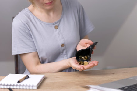 A woman pours coins, money, cents from a black purse. On the table is a notebook with blank sheets, a pen, a magnifying glass and a laptop. Poverty, economy, finance. High quality photoの写真素材
