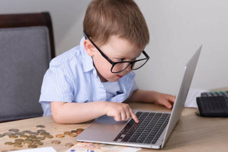 A little boy in glasses sits at a table and performs a task on a laptop. The child counts coins. Online classes. School, back to school.の写真素材