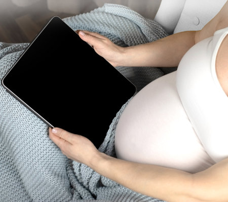 A pregnant woman sits with an electronic tablet in her hands. A pregnant woman sits working at home covered with a blue blanket. High quality photoの写真素材