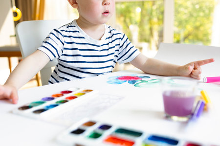A little boy sits at a table and paints with bright colors on white paper. A child learns to draw, does homework or at school, a boy in a striped T-shirt draws with his fingersの写真素材