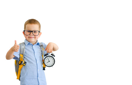 A schoolboy in a blue shirt with a backpack holds an alarm clock, shows the class with his hand. The preschooler is ready for classes, the boy is going to school, a watch in his hand, a backpack on his back. Little schoolboy with glassesの写真素材