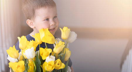 Little boy with a large bouquet of yellow tulips smiling against a sunny background. Beautiful banner with spring flowers, a gift for mothers day, birthday, holiday, a child gives flowers.の写真素材