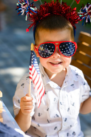 A little boy with a U.S. flag, glasses with a shiny decoration on his head is smiling. A child on a holiday, Independence Day in bright beautiful festive accessories. Happy child with glassesの写真素材
