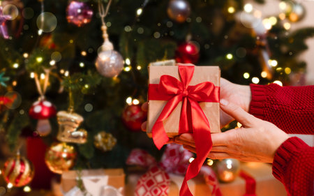 A box with a red ribbon in his hands on a blurred background with garlands. Beautiful Christmas photo. Hands with a close-up gift. New Year's Eve and Christmas Eveの写真素材