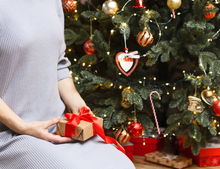 A girl sits next to a Christmas tree and holds a gift with a red bow. A box tied with a red satin ribbon, a Christmas gift, a new year's celebration, a girl with a gift.の写真素材