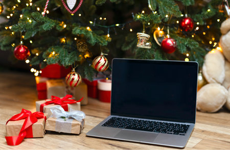 A laptop with space for a copy on a wooden background under a Christmas tree. An open laptop stands under the Christmas tree with gifts, next to them are boxes decorated with red ribbons.の写真素材