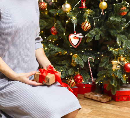 A girl in a light dress sits near the Christmas tree, holding a gift with a red ribbon. Christmas...