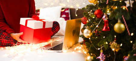 A girl sits on a bed with a large Christmas present in her hands in front of the laptop. Festive photo, communication through social networks, video call, new year, congratulations onlineの写真素材