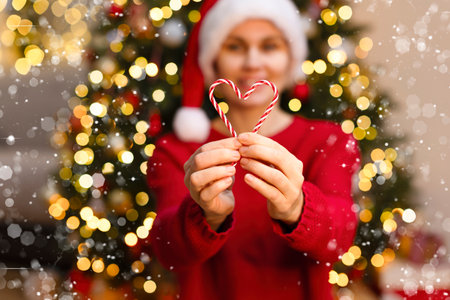 A girl in front of a Christmas tree smiles and holds candy with her heart. A defocus background with a happy woman, blurred lights, glare and radiance. Beautiful background with highlights and blur. Christmas conceptの写真素材