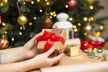 Womens hands close-up hold a packed box with red ribbon. Christmas background with a Christmas tree, a gift, blurred lights. Glare, bokeh, garland space for copy.の写真素材