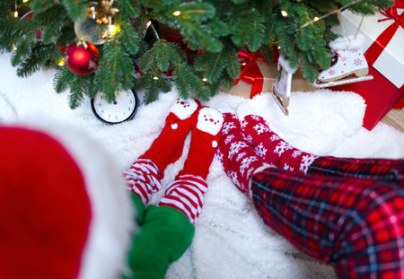 Feet in Christmas socks close-up next to the Christmas tree. A mother with a child sits on a white blanket next to a Christmas tree and gifts. Christmas, red cap, red pajamas.の写真素材