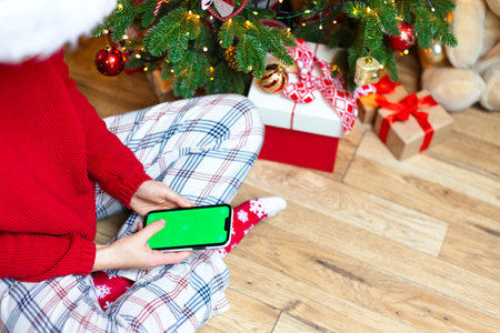 A girl in a red sweater and a Christmas cap sits near a Christmas tree with a phone. Christmas photo, holiday background, selection of gifts onlineの写真素材