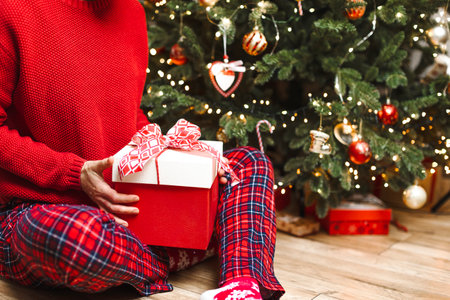 A woman opens a large gift under the Christmas tree. decorated Christmas tree with gifts, garlands, blurred lights, space for copy. Christmas photo with gifts.の写真素材