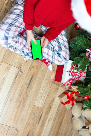 A girl in a red cap sits in front of a Christmas tree with a phone in her hands. New Years photo with a girl near a decorated Christmas tree, a smartphone with a green blank background, space for a copyの写真素材