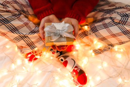 A girl sits next to a sparkling Christmas garland and holds a gift. A festive gift in the hands of a girl in a red sweater, blurred lights, glare. The concept of Christmas and New Yearの写真素材