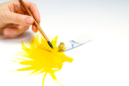 Female hand with brush drawing with yellow paints on white paper, tube with paint. The artist paints on a white canvas, a yellow painted spot or a blobの写真素材