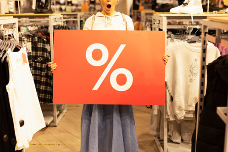 A girl in a clothing store holds a large red banner with interest in her hands. A woman has emotions of surprise and joy on her face. Discounts in the store, promotions, sale, Black Friday.の写真素材