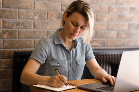 Young blonde woman in blue dress writing notes in notebook, laptop on table. Girl working in office, on wooden table in laptop office, freelancing, business womanの写真素材