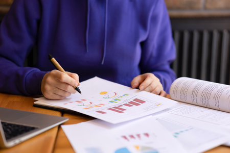Female hands making notes on sheets with graphs and data with pen. Girl with blue sweater student...