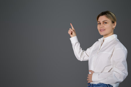 A blonde girl near a gray wall points to the space with her finger. Teacher in white office shirt pointing to empty space for copy and text, pile paste, woman smiling, whiteboardの写真素材