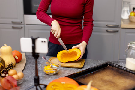 The girl cuts a pumpkin with a knife for cooking dinner and records a video on her phone. Girl shows by video link how to cut and cook a pumpkin. Thanksgiving dinner preparation.の写真素材