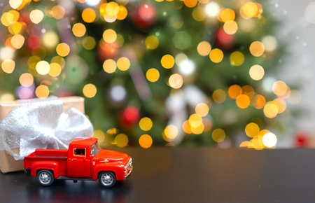 A small red car on the table with a Christmas gift and a fancy Christmas tree. Holiday card with copy space, Christmas banner. Christmas background with blurred lights and highlightsの写真素材