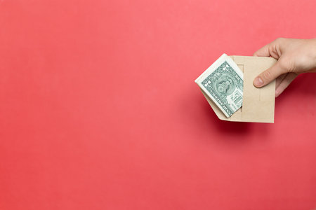 An envelope with money in hand on a red background, space for copy, text and advertising. Dollars in a brown envelope on a bright background, business, gift, payment for goods, background for advertising text. Transfer of moneyの写真素材
