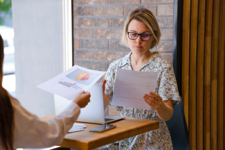 Two women working with papers and contracts in office at desk, accountant. A barefoot woman is working in an office near a window, one girl is handing over papers to another womanの写真素材