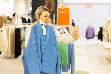 A woman in a clothing store tries on a sweater and takes a selfie. A woman shows a blue sweater for purchase by video link. Gift, new clothes, shopping.の写真素材
