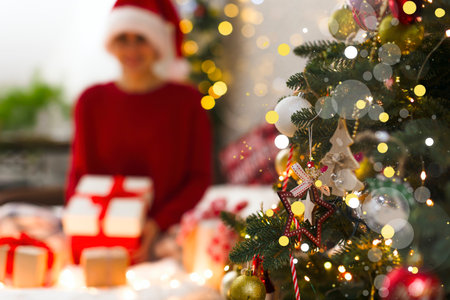 Defocus image of a girl with Christmas presents and a Christmas tree in the foreground. Christmas tree with highlights, blurred lights, gifts and a girl in a red sweater and capの写真素材
