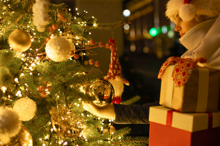 A woman with Christmas presents holds in her hand a golden ball hanging. Christmas photo, gifts, decorated Christmas tree, garlands, winter.の写真素材