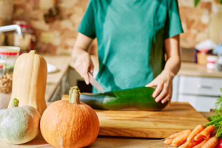 A woman in the kitchen in a green T-shirt cuts zucchini on a wooden board with a knife. Ripe orange pumpkins on a wooden kitchen table, space for a copy.の写真素材