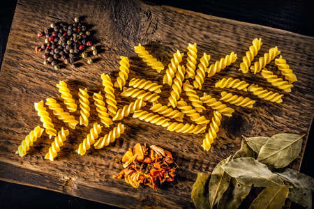 Background Italian spiral paste, bay leaves, spices and multicolored round peppers lie on a textured wooden surface.の写真素材