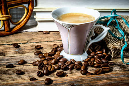 Cup of coffee with froth and scattered beans on a wooden table on a background of booksの写真素材
