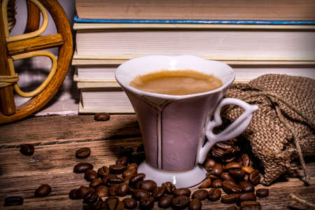 Cup of coffee with froth and scattered beans on a wooden table on a background of booksの写真素材
