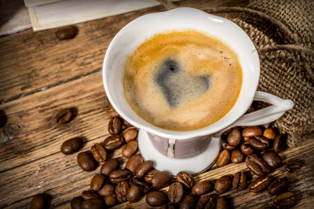Cup of coffee with a heart painted in foam and beans scattered from a bag on a wooden tableの写真素材
