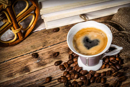 Cup of coffee with a heart painted in foam and beans scattered from a bag on a wooden table on a background of booksの写真素材